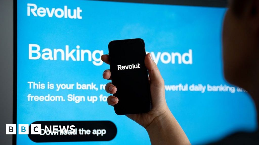 A person holding a smartphone displaying the Revolut app logo, in front of a display showing a bright blue Revolut advertisement.